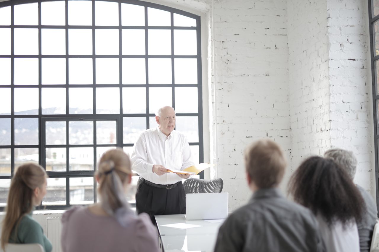 Home Multiracial coworkers in casual wear listening to aged gray haired coach at corporate group meeting in modern contemporary office boardroom